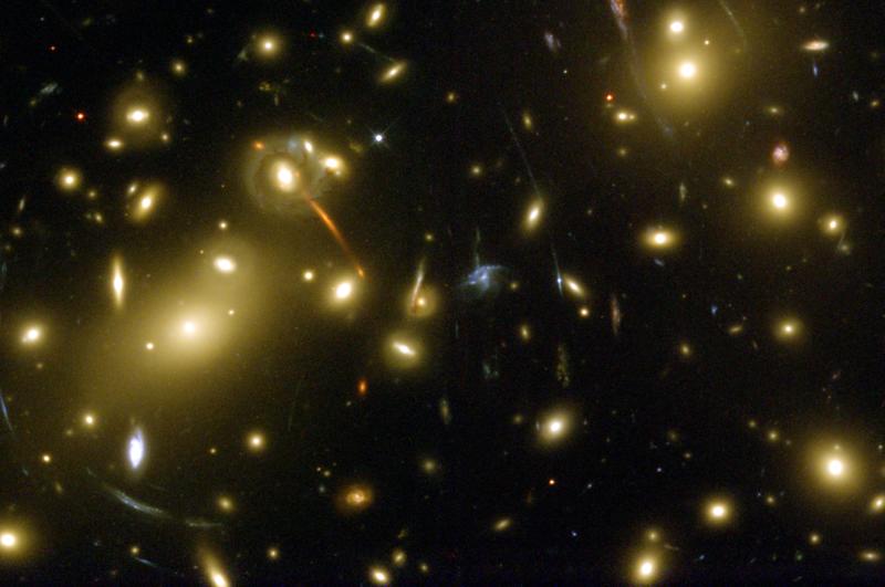 Hubble Space Telescope image of the galaxy cluster Abell 2218. In the foreground we observe a large number of galaxies which make up the cluster (the larger galaxies, with typically gold colors representative of early-type (elliptical) galaxies). The fainter galaxies which appear as elongated streaks are those located in the background much further away from us. Most of the streaks are centered around the center of the foreground cluster, because it is its mass which distorts the background light as it passes towards us.