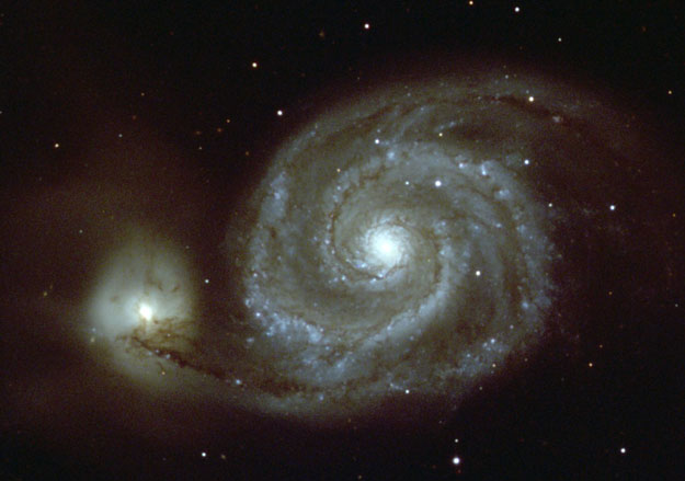 Image of M51 (aka the Whirlpool galaxy), a large face-on spiral galaxy with clearly defined spiral structure and extended spiral arms looping around the central bulge. Note the presence of a smaller yellow galaxy passing behind M51 and tugging on the stars along one spiral arm.
