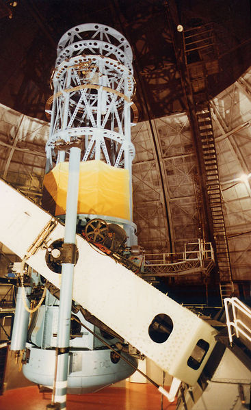 Photograph of the 100-inch telescope at Mount Wilson. The telescope is a large impressive cylinder of metal supports for lenses, cameras, and other components. It is mounted on a mechanical system that allows it to be rotated in place to follow the movement of an object across the sky, and surrounded by a large dome to protect it from the elements.