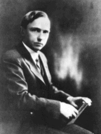 Photograph of Harlow Shapley, shown seated while wearing a suit and tie and holding a book in his hands.