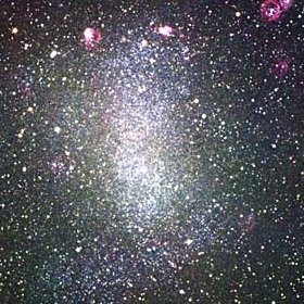 Telescope image of NGC 6822, showing an elongated bright white structure and patches of red and white light embedded in a diffuse cloud of white light.