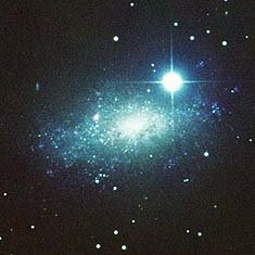 Telescope image of IC 5152, showing white and greenish light (this is because of the particular filters used to image the galaxy) in a patchy elongated blob. A bright foreground star shines strongly in the image as well.