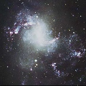 Telescope image of NGC 1313, showing bright white patches and arcs of light and a dense white core, with disordered diffuse light and smaller white clumps surrounding it.